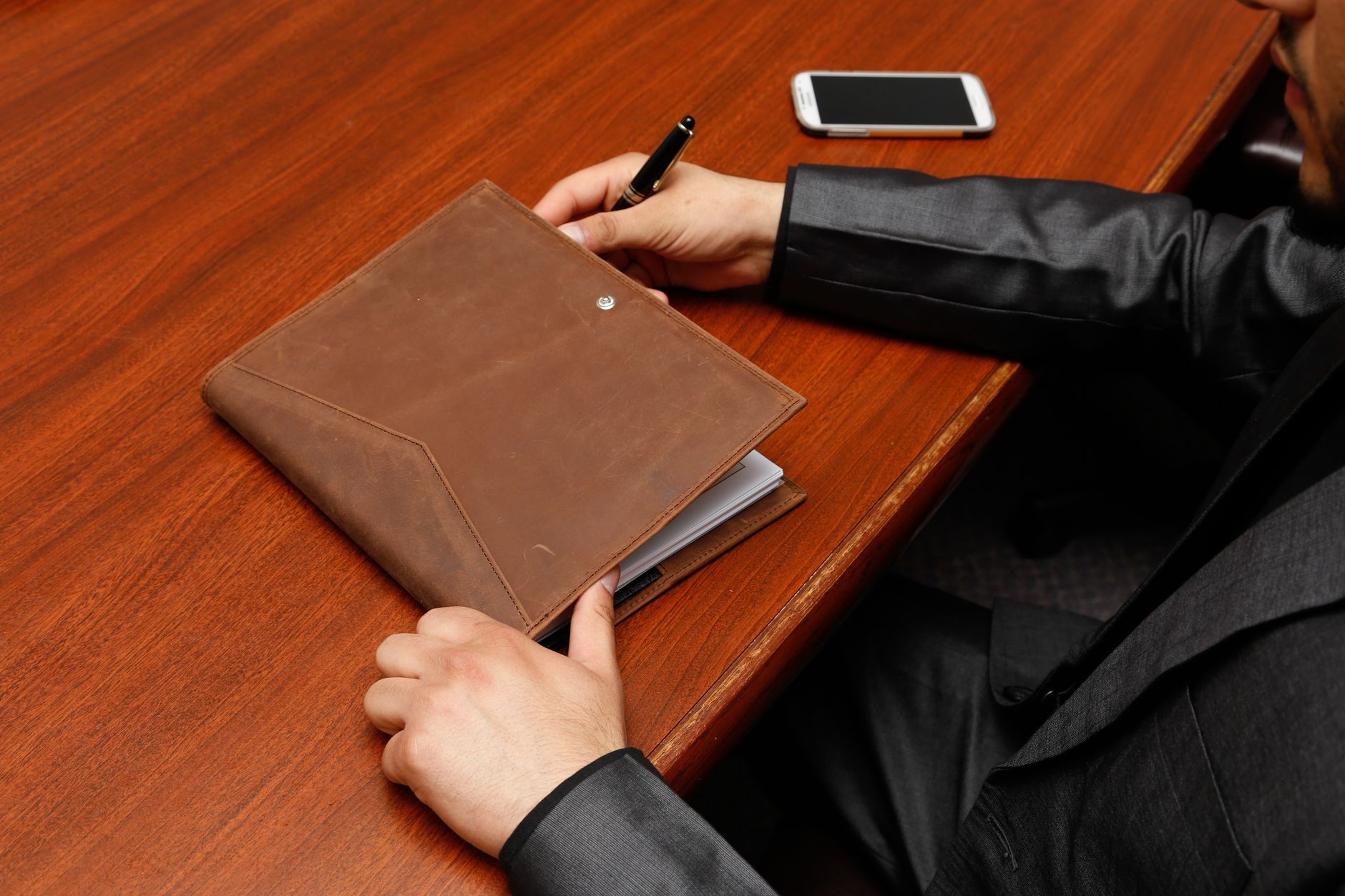 A person in business attire writing notes into a leather notebook at a wooden desk, with a phone nearby.